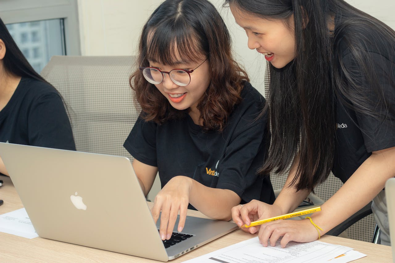 Two women smiling and working together on a laptop, embodying teamwork and collaboration in an office.